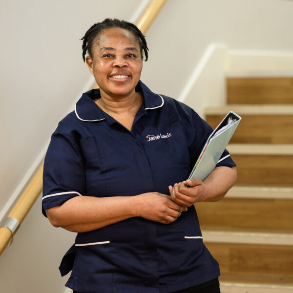 Female nurse wearing Jane Lewis uniform holding a folder standing in front of some stairs in a service