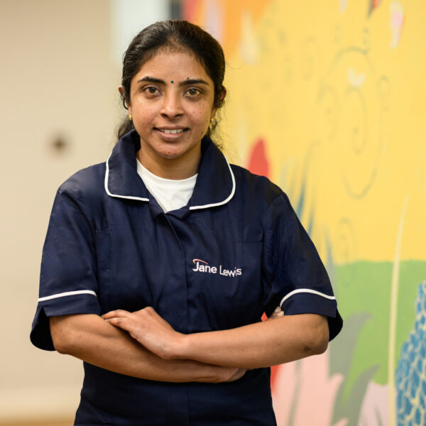 Female nurse, wearing a Jane Lewis navy blue, with her uniform arms folded, standing in a bright service hallway