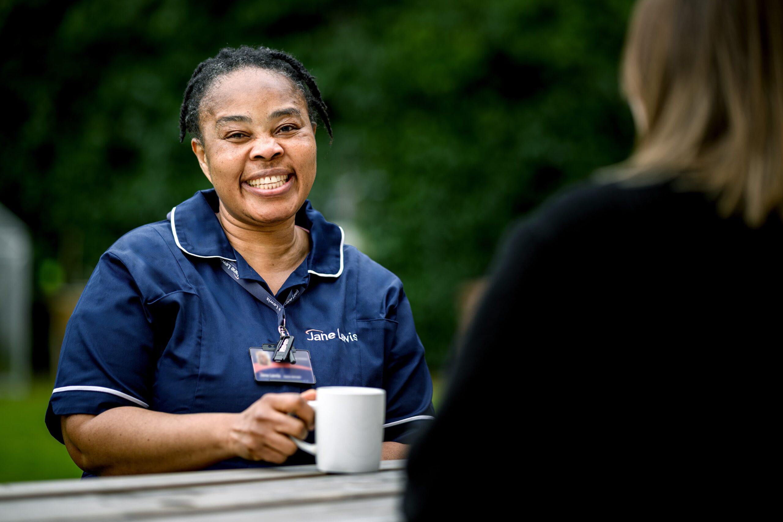 Nurse in a Jane Lewis uniform sitting on a picnic bench with a cup of tea
