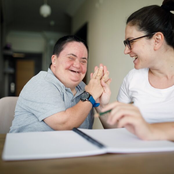 Carer supporting resident in a supported living facility, sat at a dining table with a notebook
