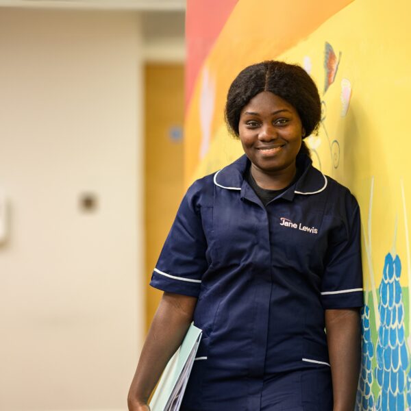 Female nurse in Jane Lewis uniform leaning against wall in service corridor with folder in hand
