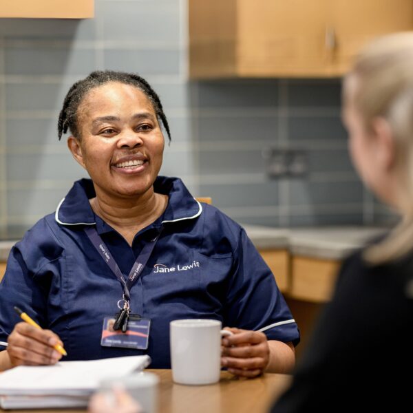 Female nurse in Jane Lewis uniform sat at kitchen table with service user, taking notes