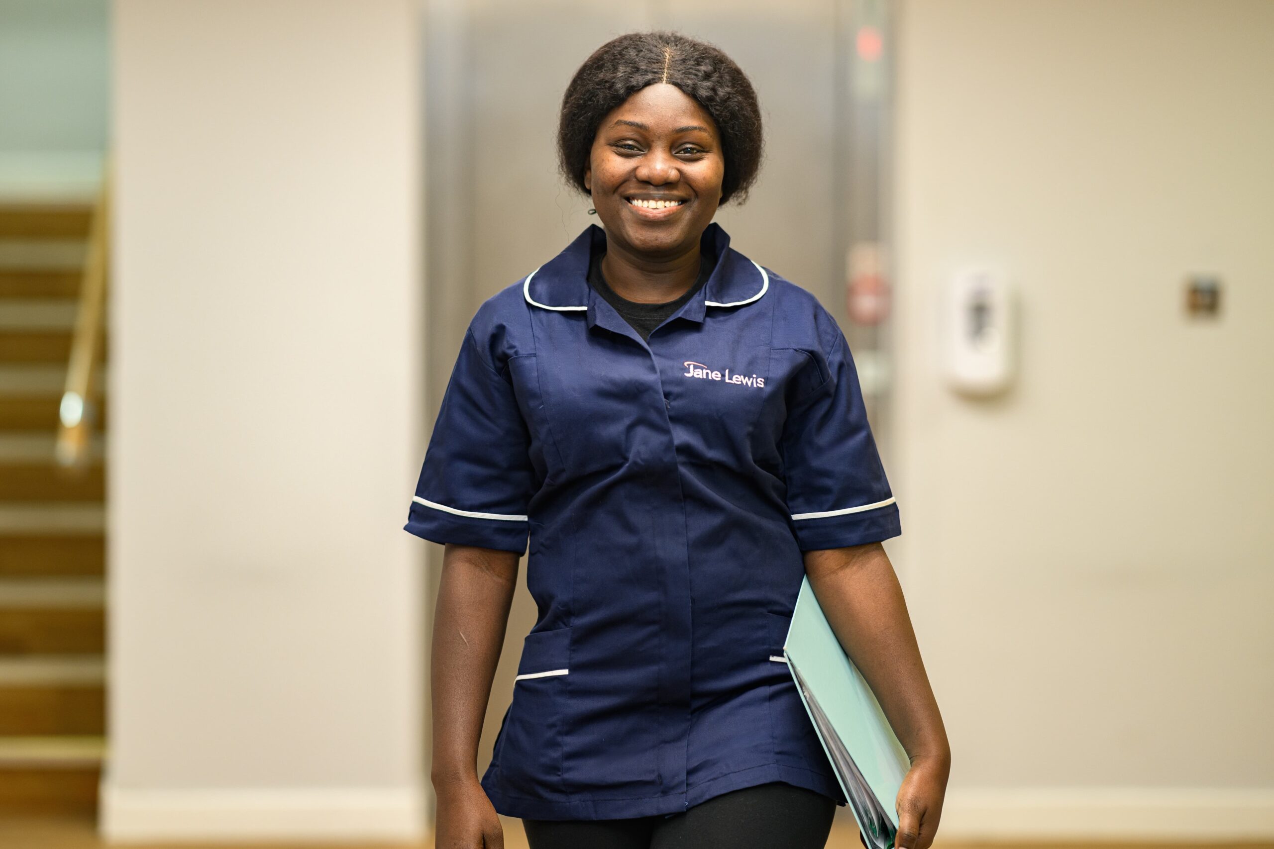 Female nurse in Jane Lewis uniform walking through the corridor in a service with a pale blue folder in hand