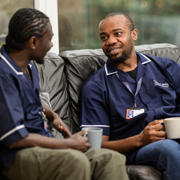 Two male Jane Lewis nurses sat on a sofa enjoying a hot drink on their break